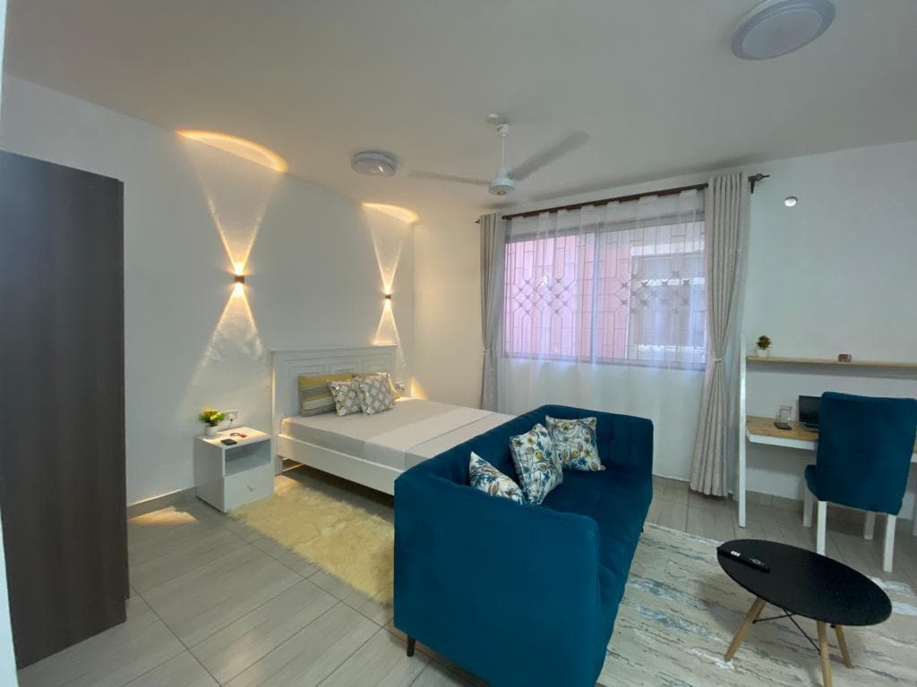 A well-lit studio space features a comfortable bed with decorative pillows and a cozy seating area with a blue sofa. Natural light filters through the sheer curtains, and a small work desk with a chair is positioned nearby, contributing to the room's functional layout.