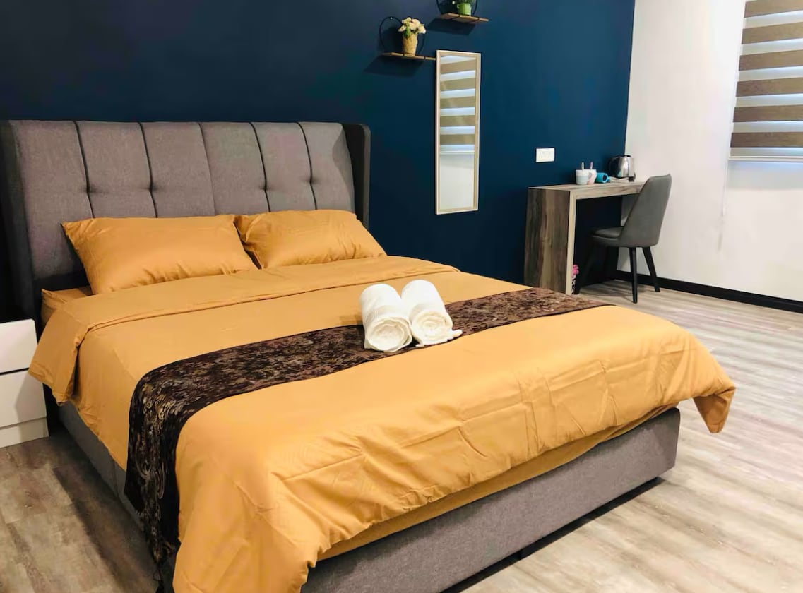 A cozy sleeping area features a spacious bed with soft, golden linens and a decorative dark brown throw. Two towels are neatly arranged at the foot of the bed. A small desk with a chair and a mirrored shelf are visible against the contrasting blue wall.