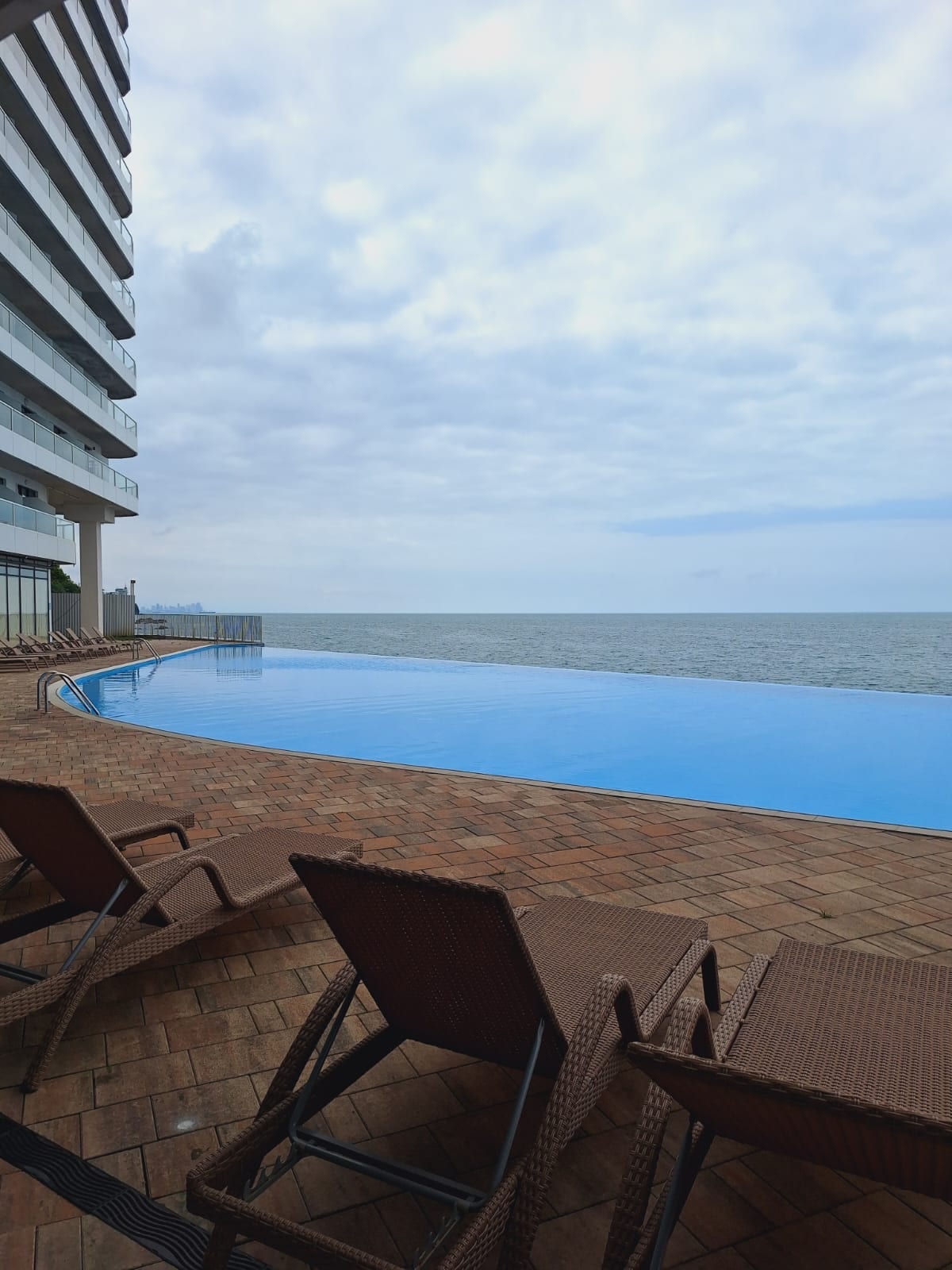 An infinity pool is situated beside the ocean, displaying a seamless blend of water and sky. Several sun loungers are arranged along the poolside, offering a relaxing space to enjoy the view. The tranquil atmosphere is enhanced by the gentle waves in the distance.