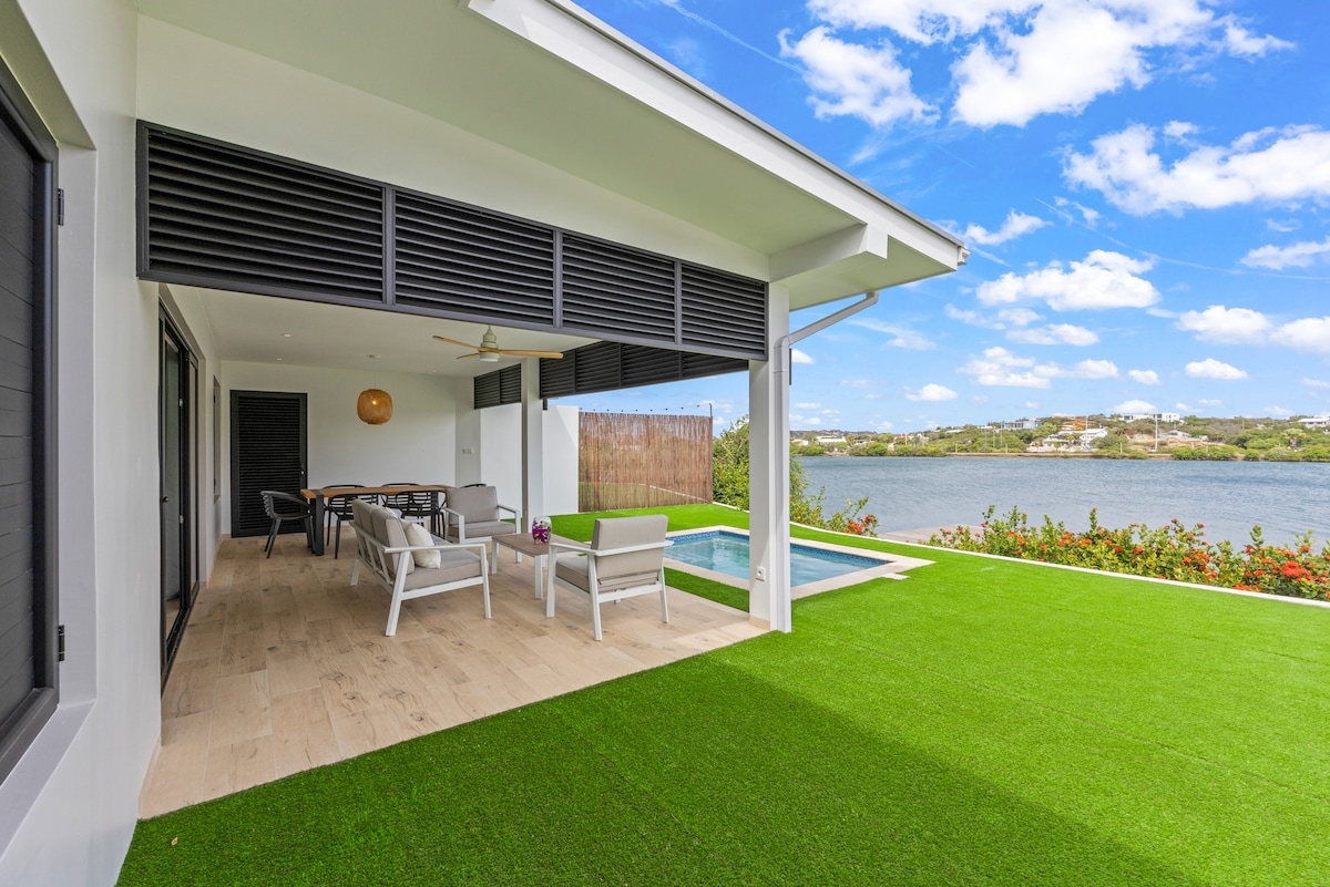 An outdoor living space extends along the water's edge, featuring a dining table with seating for six and a private pool. Lush green grass surrounds the area, while panoramic views of the waterfront and sunny blue skies enhance the peaceful atmosphere.