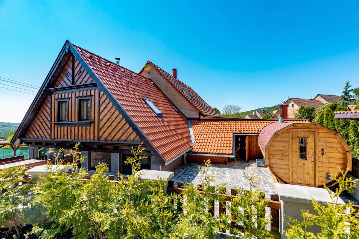 A charming exterior of a two-story home is showcased, featuring a unique sloped roof with vibrant red tiles. Wood accents enhance the facade, while a circular wooden sauna is visible to the side. Lush greenery surrounds the property, contributing to a serene outdoor environment.
