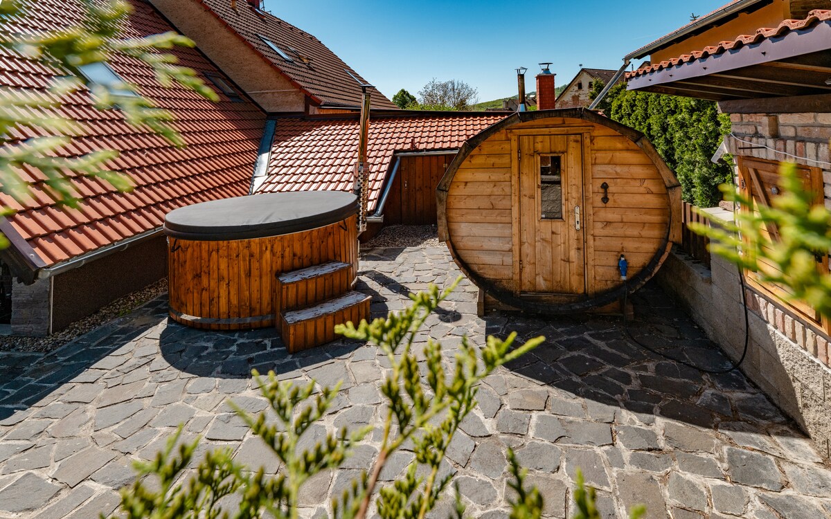 An outdoor area features a wooden hot tub, accompanied by a sauna structure. The space is paved with stone, surrounded by a low wooden railing. Nearby rooftops are visible, providing a rustic backdrop under a clear blue sky.