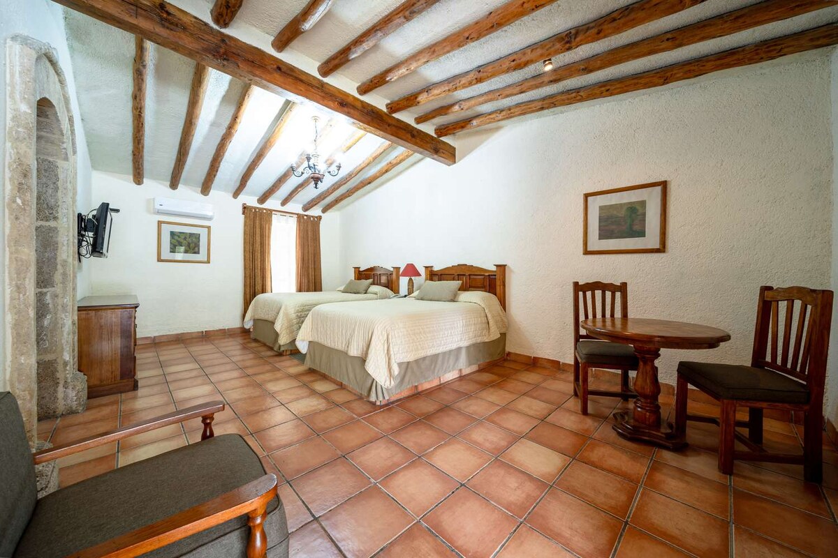 The room features a high ceiling with exposed wooden beams, contributing to an airy feel. Two spacious beds are positioned opposite a wooden table and chairs. A small television is mounted on a stone wall, and large windows are adorned with curtains, allowing natural light.