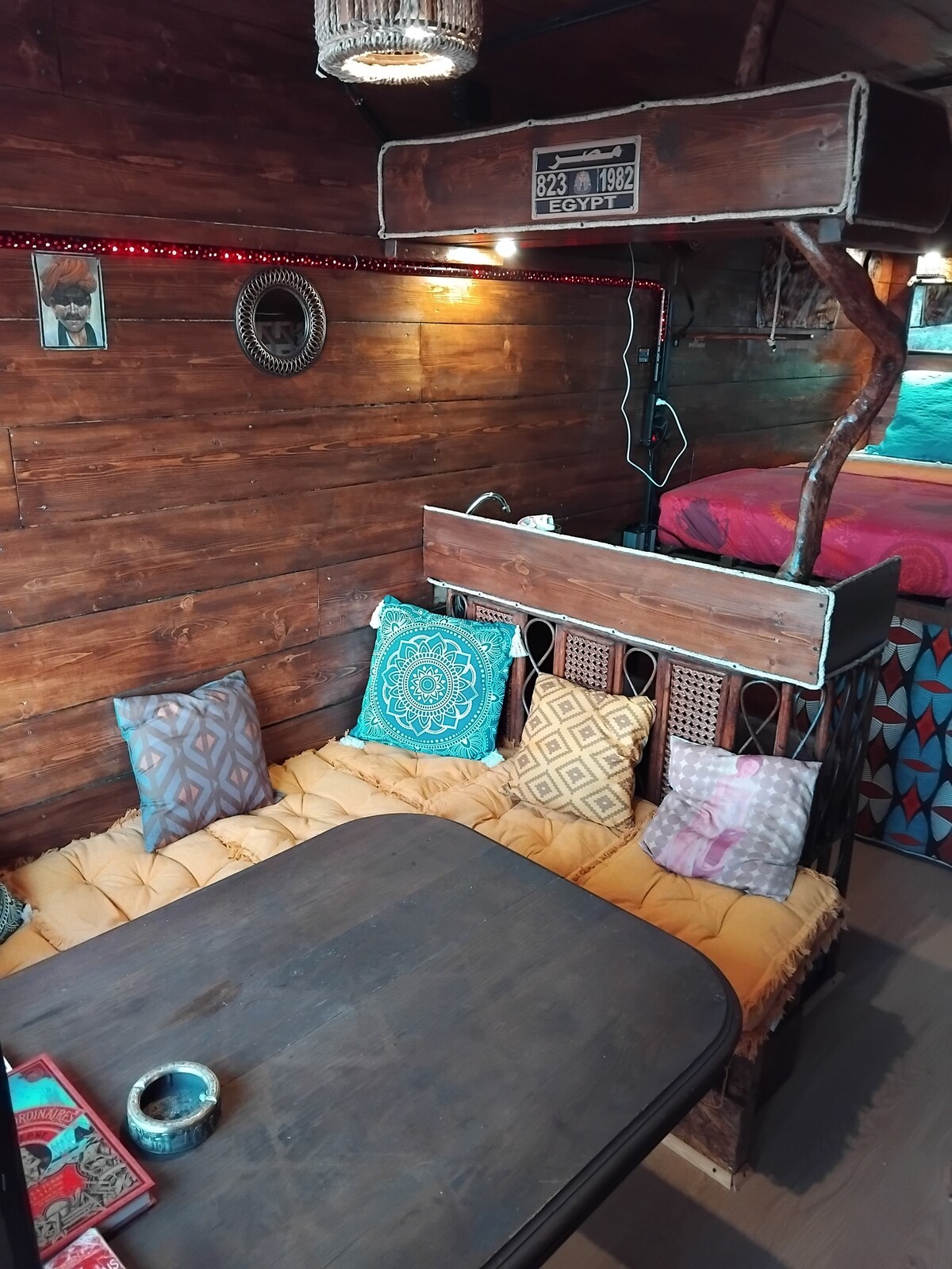 A cozy seating area is featured, with a wooden table and a comfortable bench adorned with patterned cushions. The walls are constructed of rich wood, and decorative elements enhance the warm ambiance. Above, a sleeping loft is visible, framed by natural wood accents.