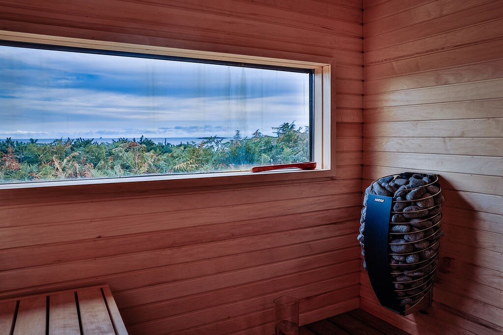 A wooden sauna interior is showcased, featuring a large window that offers views of surrounding greenery. A stone heater is positioned on one wall, while a wooden bench runs along the opposite side, creating a serene space for relaxation.