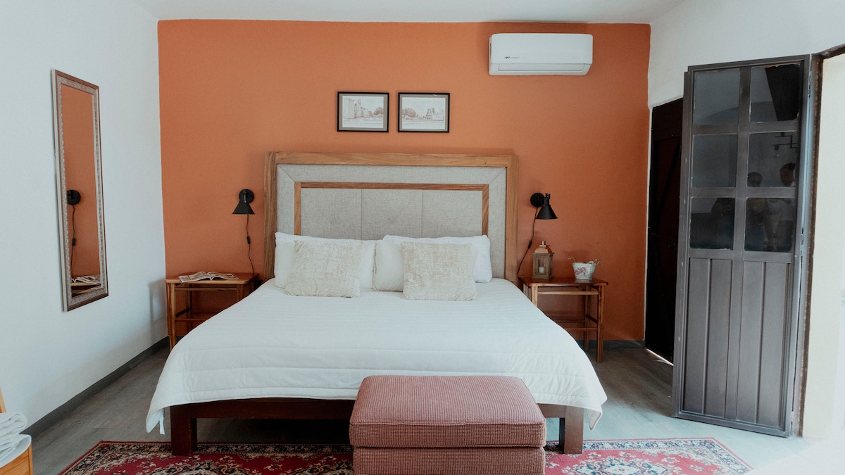 A spacious sleeping area features a large bed centered against an orange wall. Two lamps frame the bed, providing subtle lighting. A decorative ottoman is positioned at the foot of the bed, and a mirror reflects the room's warm tones. A door leads outside.
