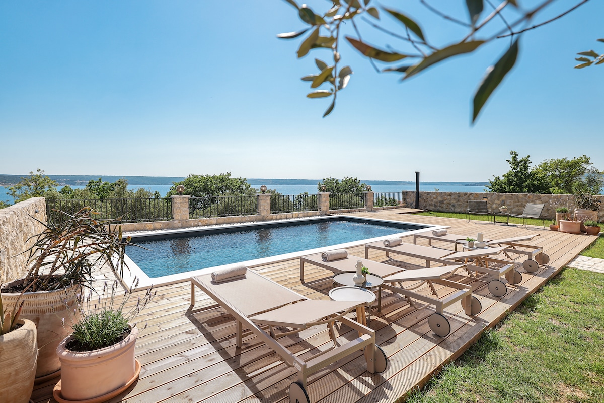 A serene outdoor area features a large swimming pool surrounded by wooden decking and several lounge chairs. Lush greenery and plants enhance the space, while a clear view of the sea is visible in the background, under a bright blue sky.