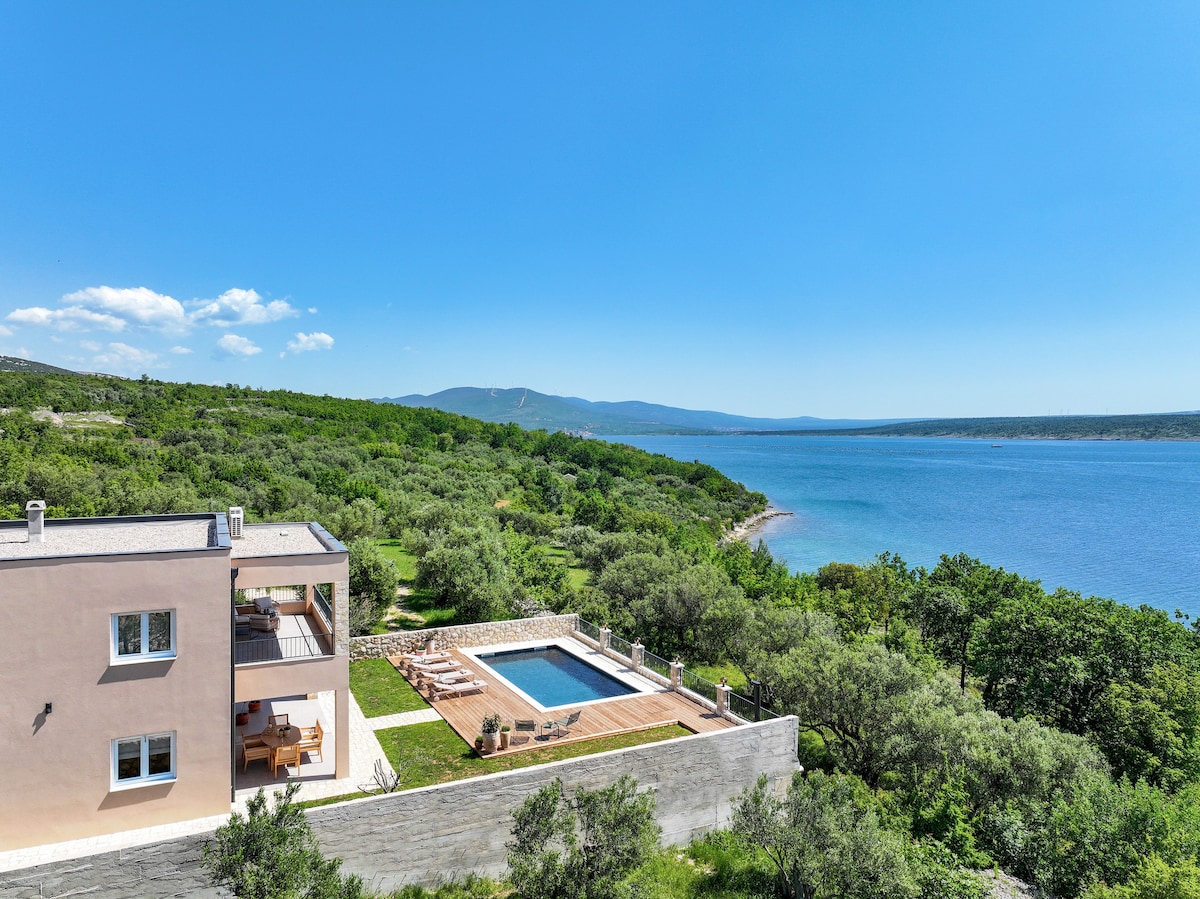 A spacious villa is set on a hillside, offering an expansive view of the coastline. A large swimming pool is surrounded by wooden decking, and lush greenery frames the property, creating an inviting outdoor space. The clear blue sky enhances the serene atmosphere.
