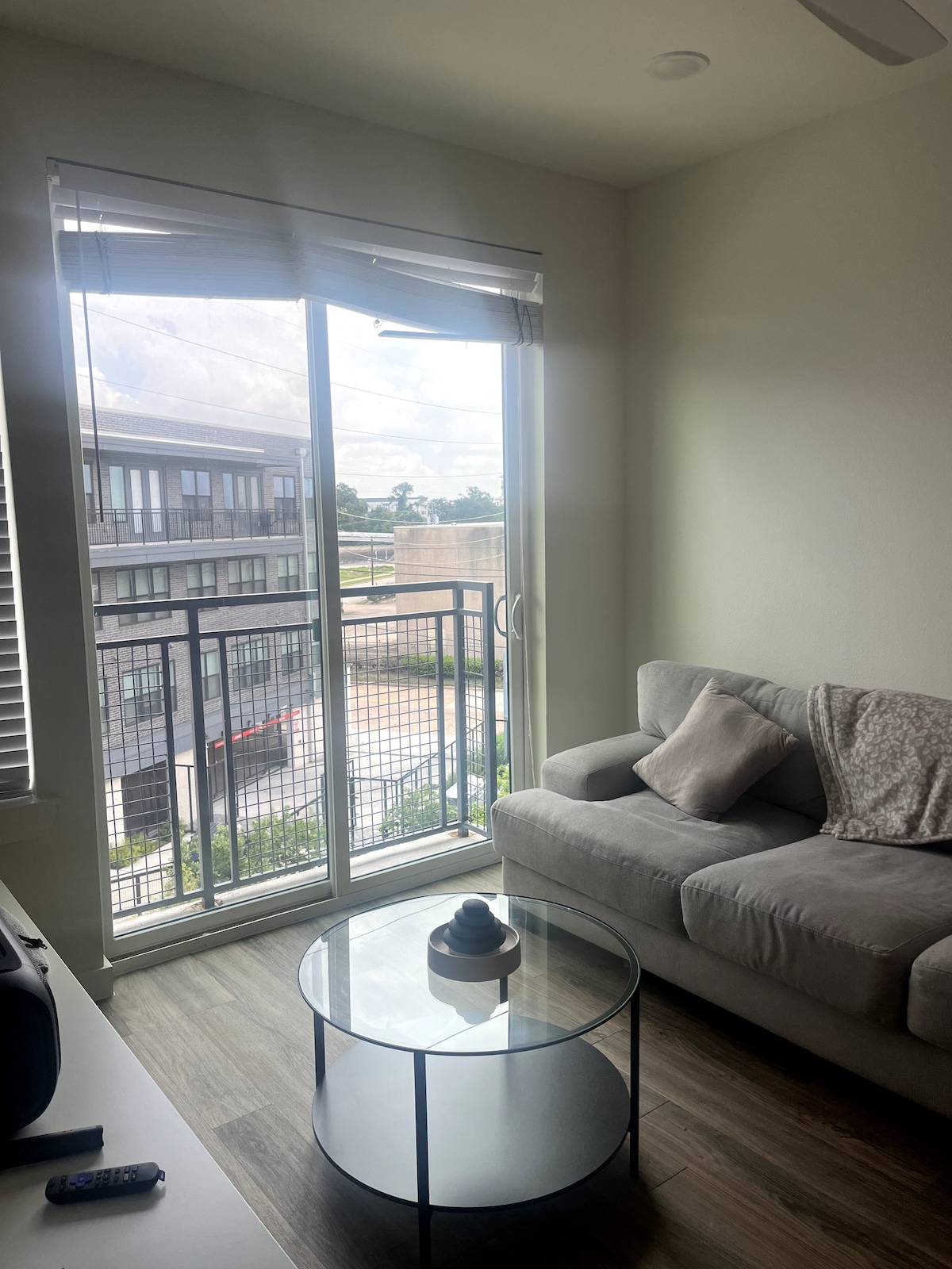 A bright living area features a light gray sofa placed near a glass coffee table. Large windows offer an unobstructed view of the outside. Natural light fills the space, accentuating the modern design of the room.