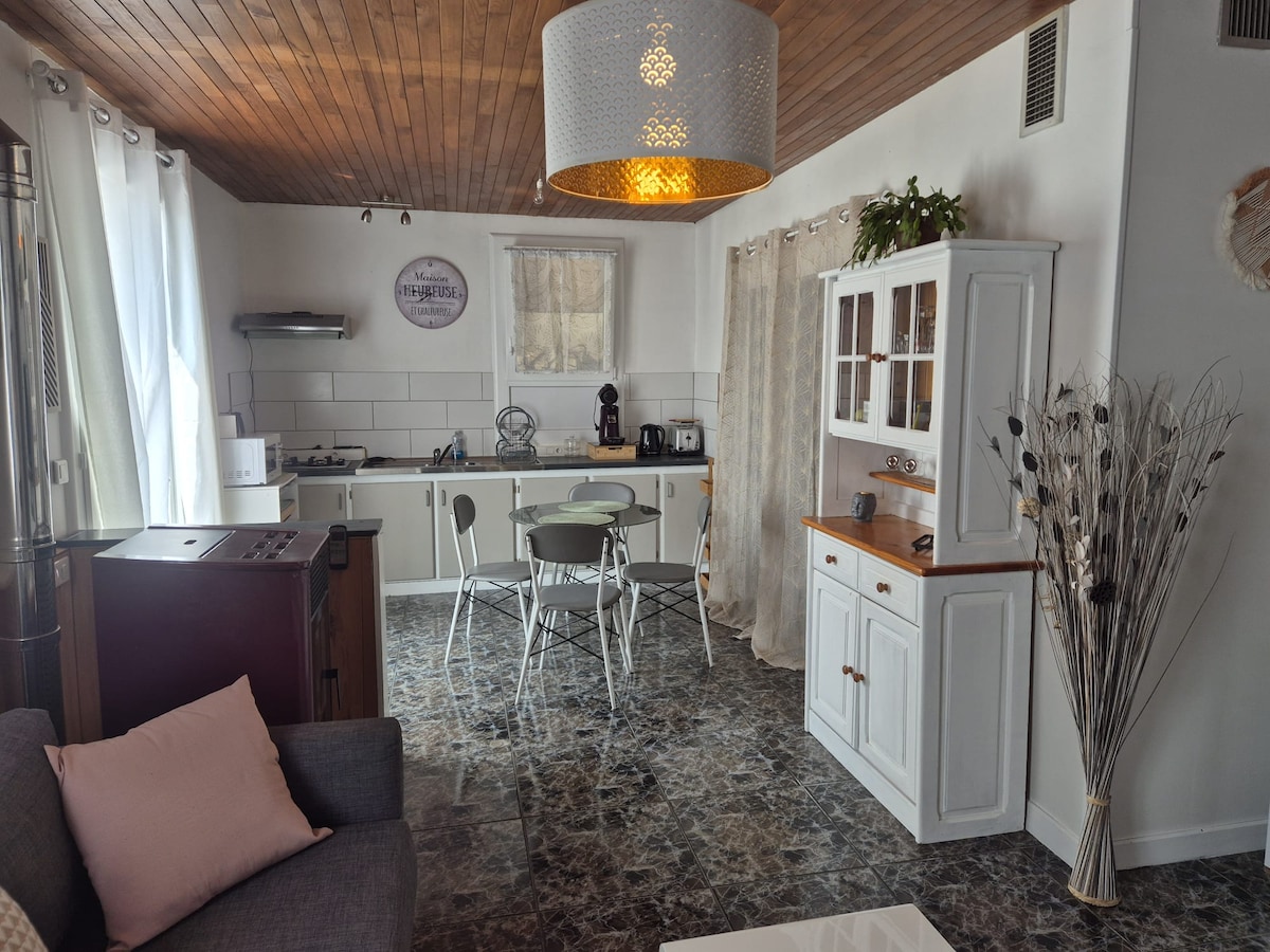 The living space features a modern kitchen with white cabinetry and a dark countertop. A dining area is set with four chairs around a round table. Soft natural light enters through large windows, accentuating the textured ceiling and warm wooden elements.