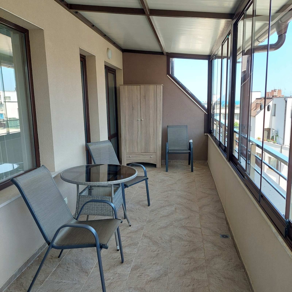 A sunlit balcony is furnished with a round glass table and two chairs. A wooden cabinet is placed against the wall, providing additional storage. Large windows flood the space with natural light while offering a view of the surrounding area.
