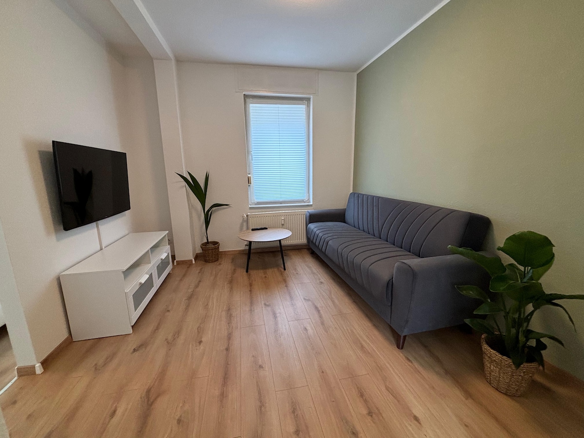 A living area showcases a light green accent wall and features a cozy couch with a modern design, paired with a small round coffee table. A flat-screen TV is mounted on the wall, and a tall plant complements the space. Natural light enters through a window with horizontal blinds.