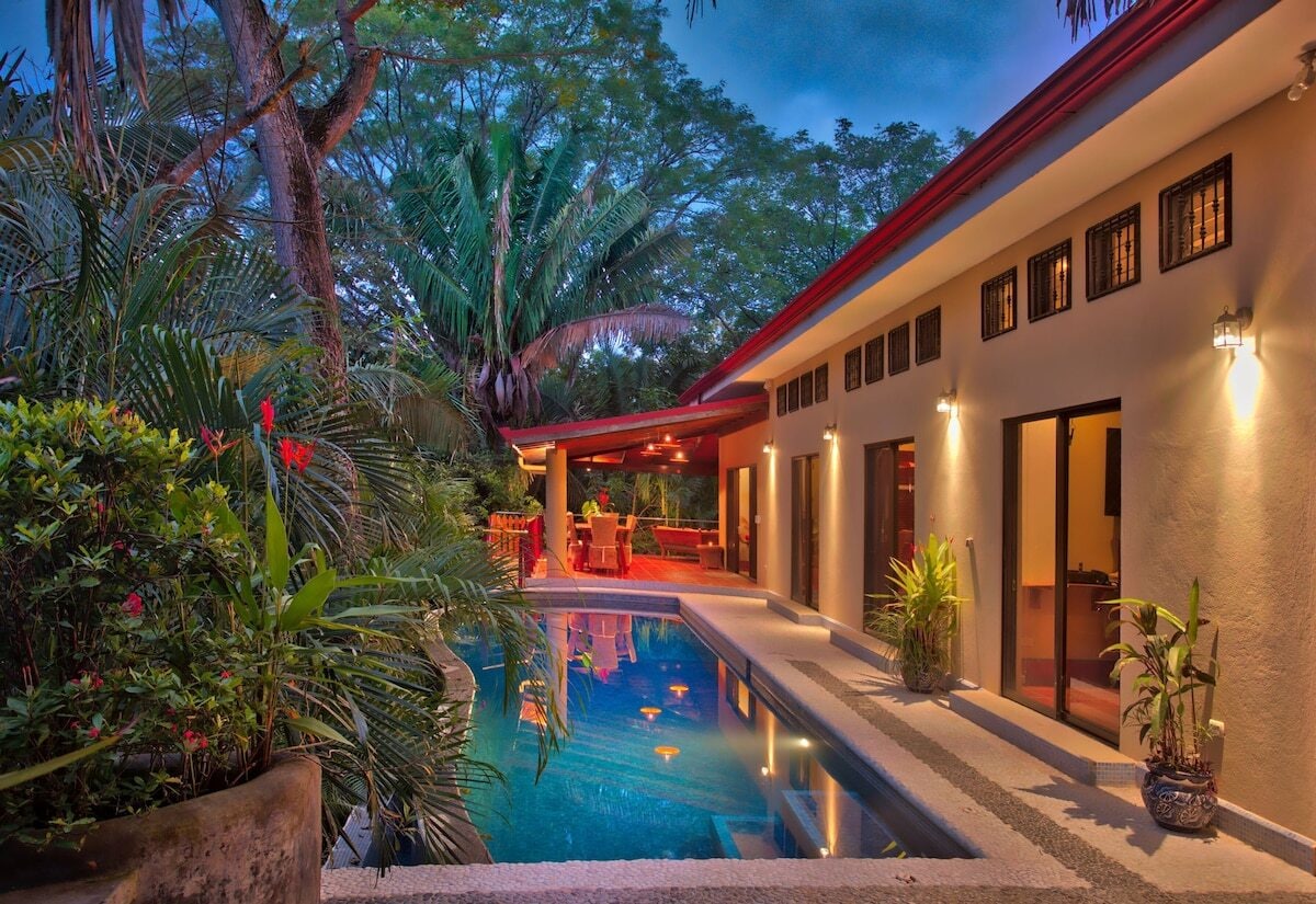 A serene outdoor space features a tranquil pool surrounded by lush tropical foliage. The home's exterior, highlighted by soft lighting, showcases large windows and inviting seating areas. This setting offers a peaceful retreat nestled in the natural landscape.