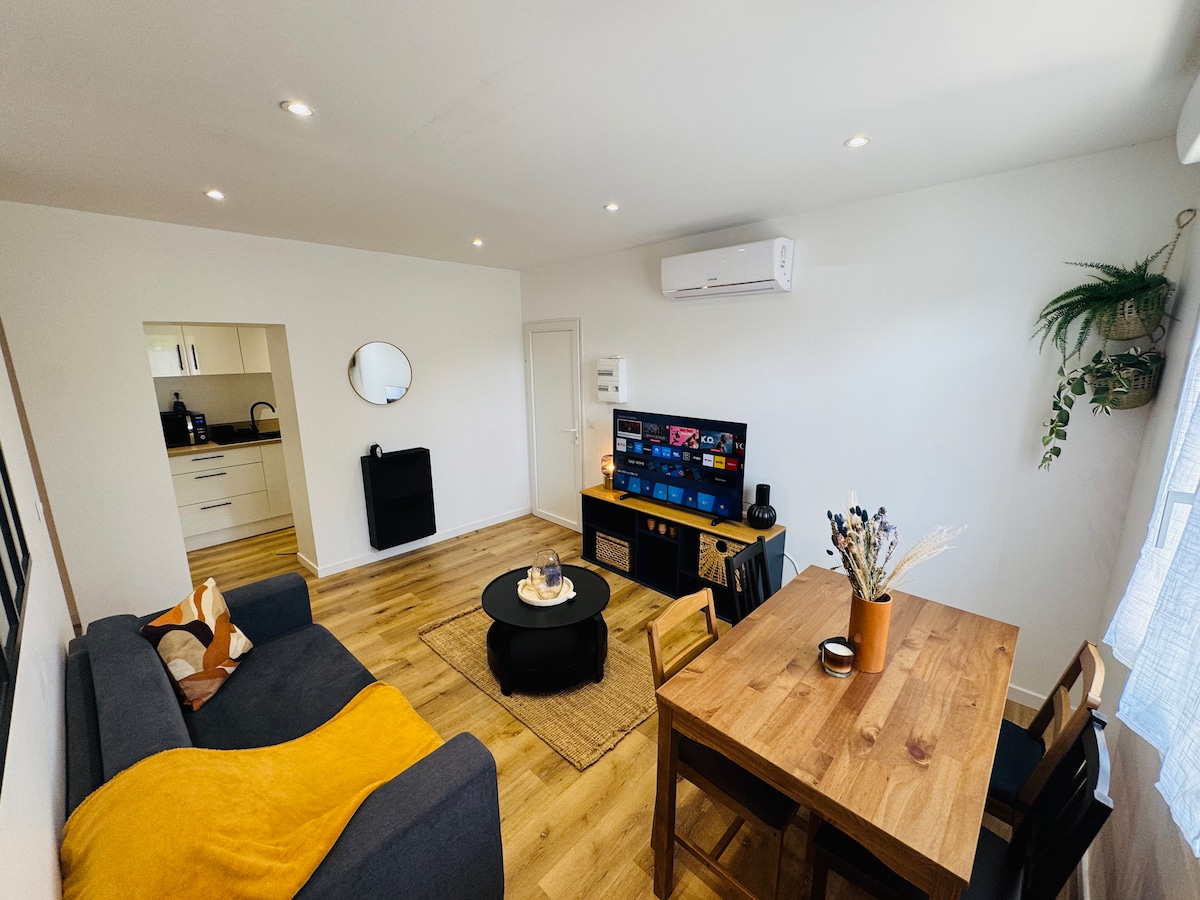 A modern living area features a comfortable gray sofa adorned with an orange throw, a circular black coffee table, and a wooden dining table with chairs. A flat-screen TV is positioned on a media console, with ambient light provided by recessed ceiling fixtures.