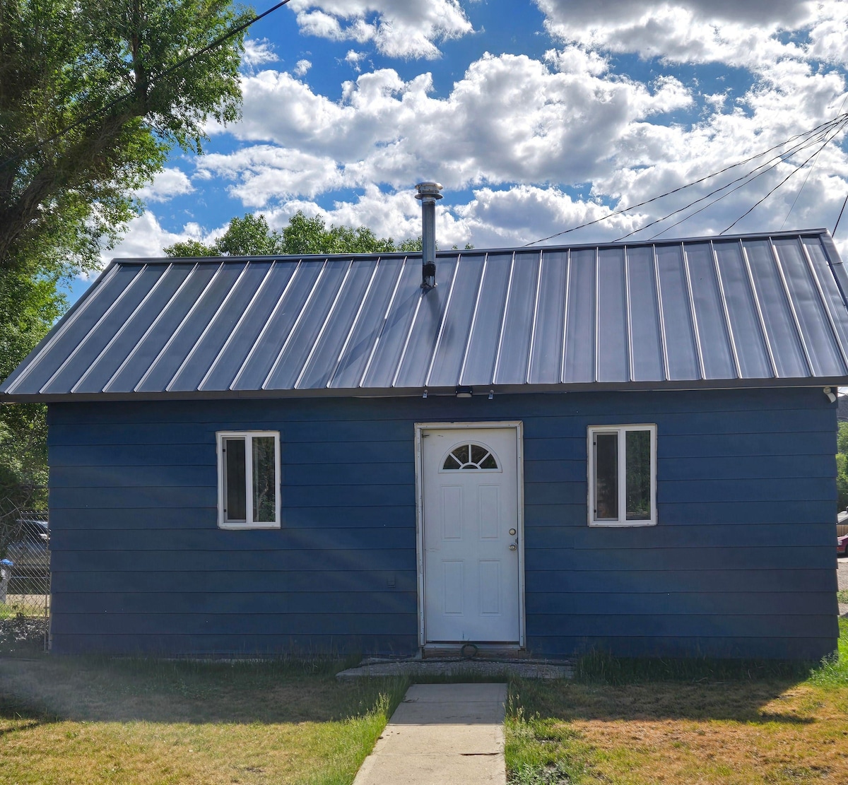 The exterior of a blue one-story home is presented, featuring a modern metal roof and a centrally positioned front door. Two small windows are visible on either side of the entrance. A pathway leads to the door, with grass surrounding the foundation.