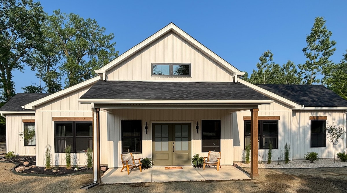 The home's exterior showcases a charming farmhouse design with light-colored siding and dark roofing. Two wooden rocking chairs are positioned on the covered porch, while well-maintained greenery frames the entrance. Large windows allow natural light to illuminate the interior.