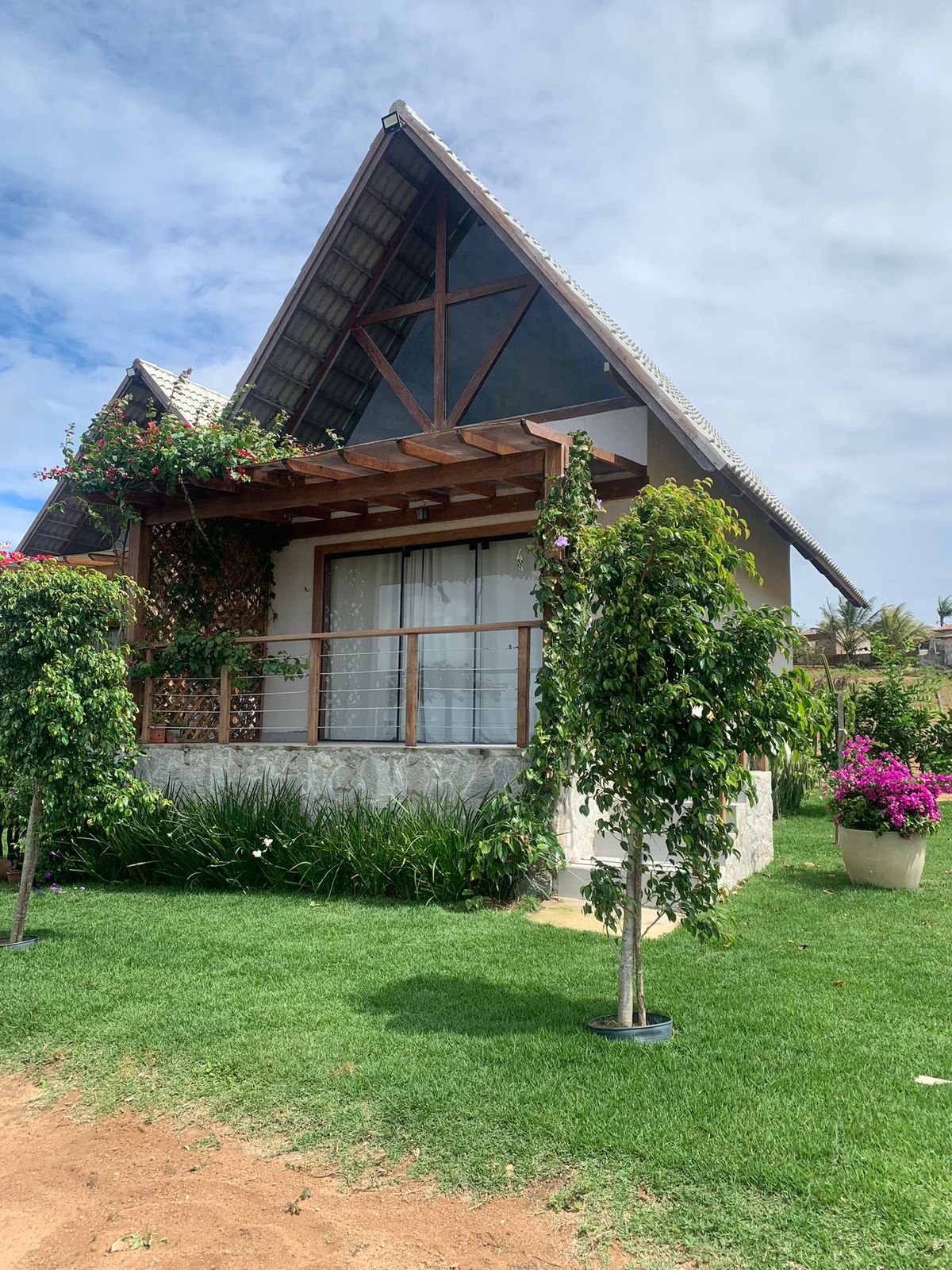The exterior of a modern chalet is depicted, featuring a sloped roof and a wooden balcony adorned with vibrant greenery. Lush grass surrounds the entrance, complemented by potted plants and trellises, creating a tranquil and inviting outdoor space.