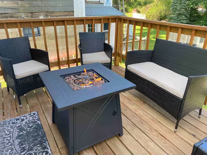 A deck area is showcased with a square fire table at the center, surrounded by two armchairs and a two-seat sofa, all featuring dark wicker surfaces and light cushions. The wooden deck, with a railing, provides a view of the green yard beyond.