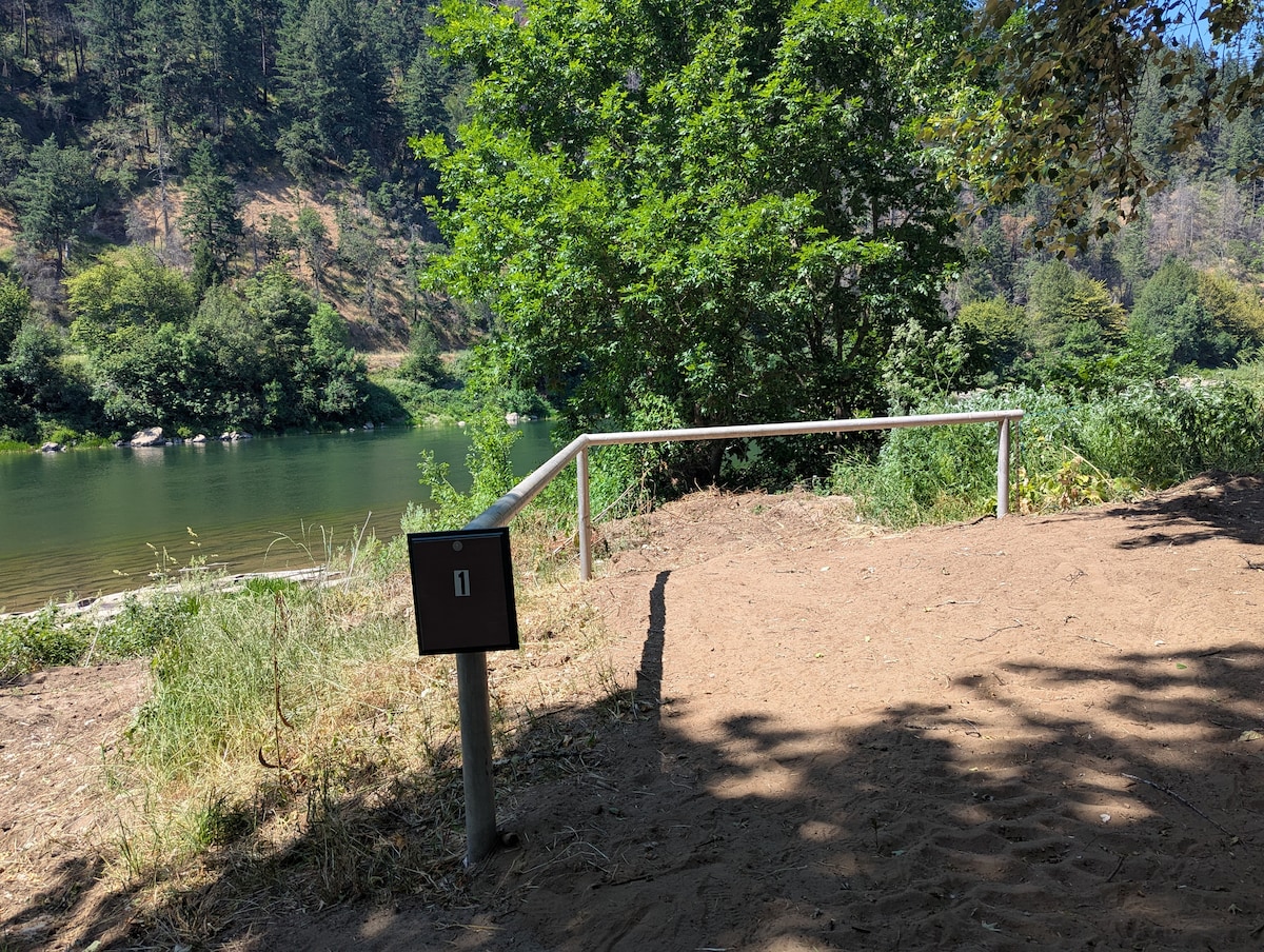 A sandy campsite area is visible near the riverbank, with a metal railing providing support to guests. A wooden sign labeled '1' marks the site, surrounded by lush greenery and trees, while the calm waters of the river reflect the landscape.