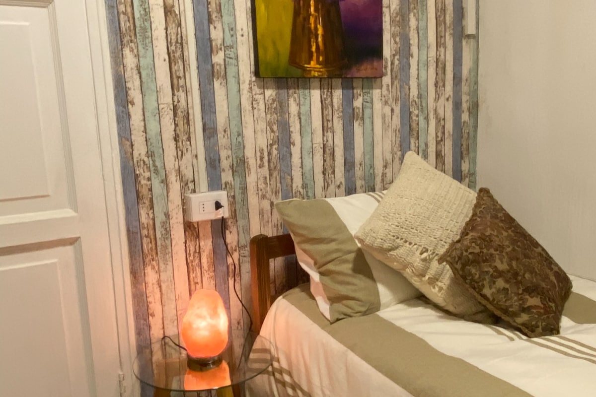 A comfortable sleeping area is complemented by a bedside table featuring a glowing salt lamp. Textured pillows are arranged on the bed, which is accompanied by a decorative piece on the wall. Subtle blue and white striped wallpaper creates a serene backdrop.