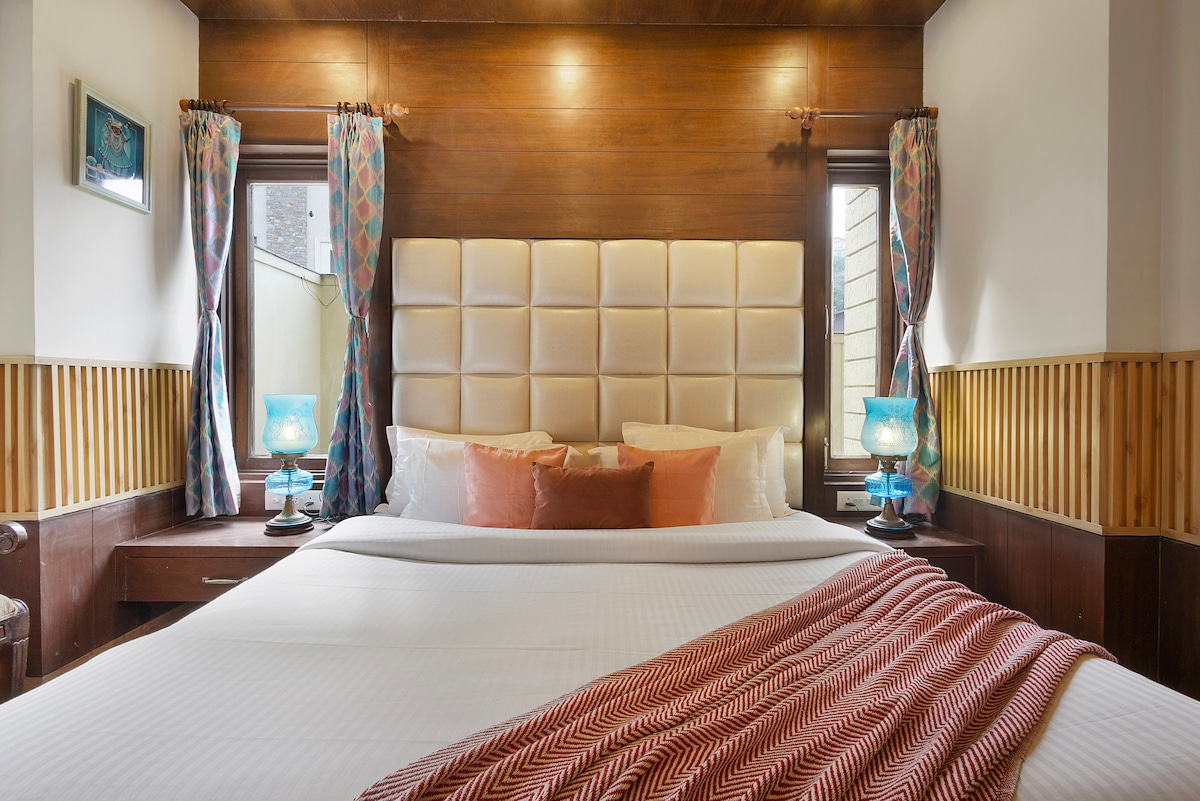 A well-appointed bedroom features a spacious bed with a tufted headboard and decorative pillows. Soft lighting is provided by bedside lamps. Colorful curtains frame the windows, and polished wood accents contribute to a warm ambiance.