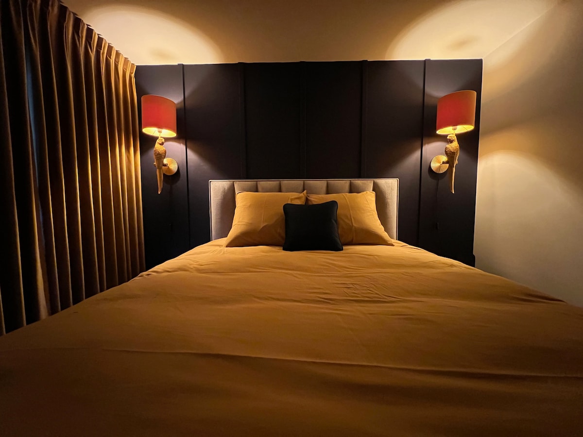 A spacious bed is centered against a dark, padded headboard, flanked by stylish wall sconces that emit a soft glow. Rich, golden curtains frame the window, enhancing the cozy ambiance of the sleeping area.
