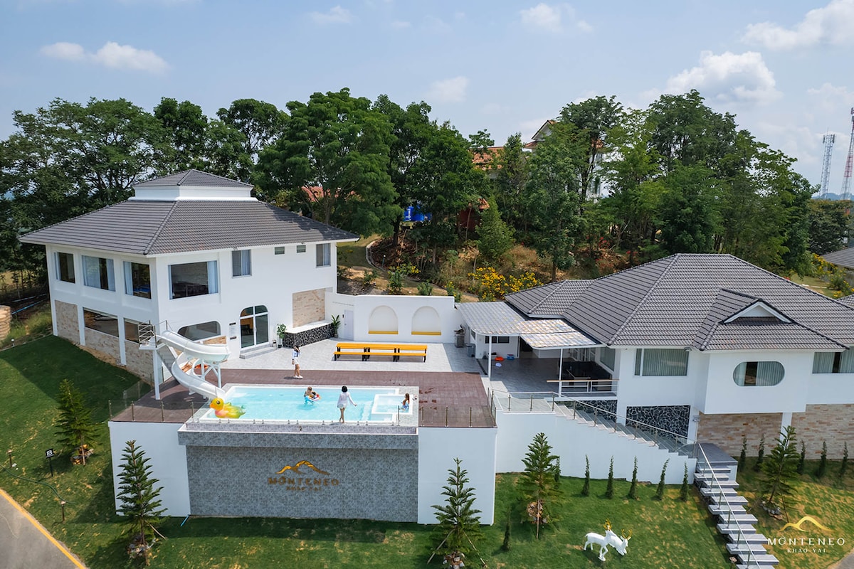 An aerial view of a modern villa surrounded by lush greenery, featuring a private swimming pool and a large deck area. The exterior showcases a combination of white walls and natural stone, with a spacious terrace furnished with seating. The property is designed for relaxation and outdoor enjoyment.