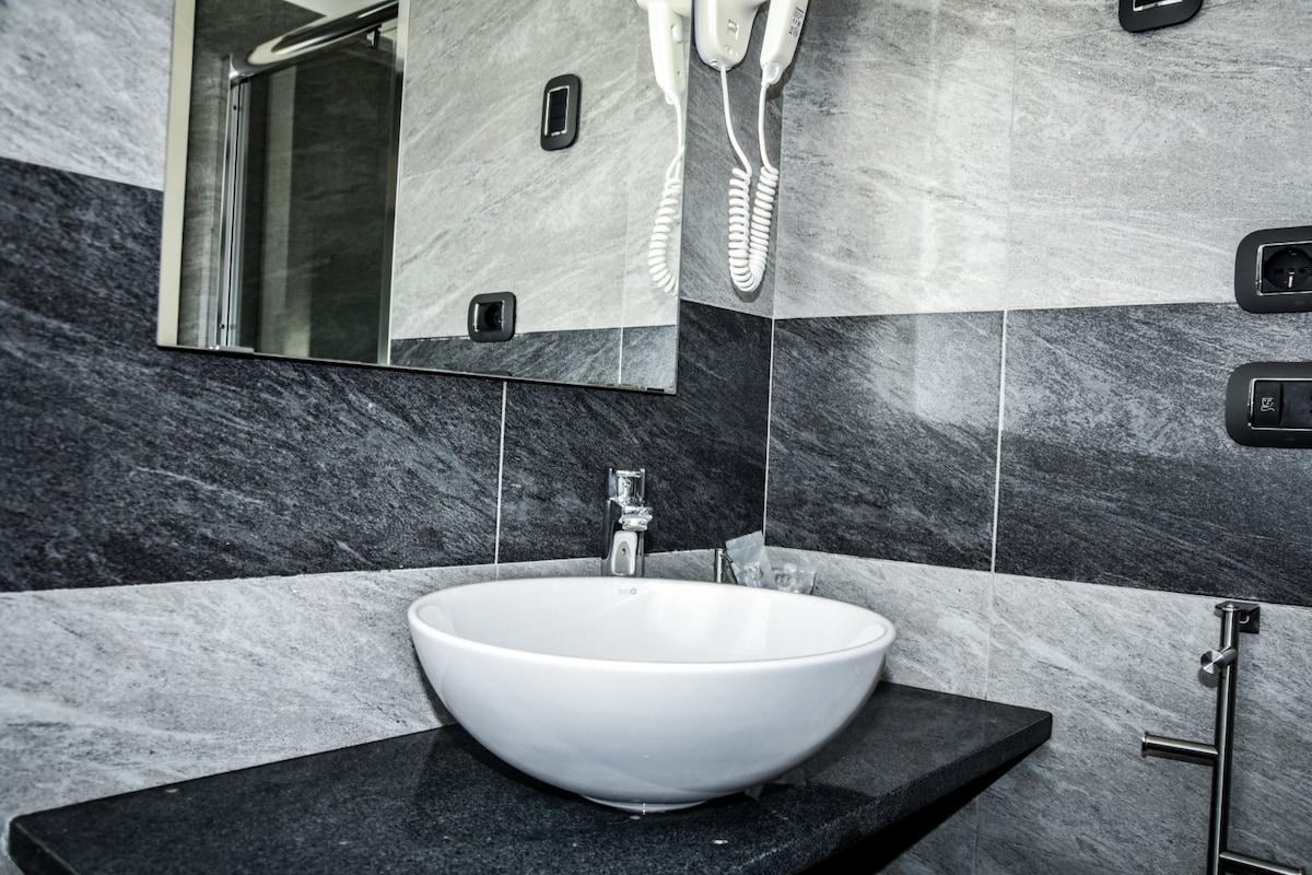 A modern bathroom features a sleek, circular sink resting on a dark granite countertop. The walls are adorned with contrasting grey tiles, and a mirror reflects the space. An overhead hairdryer is mounted nearby, contributing to a functional design.