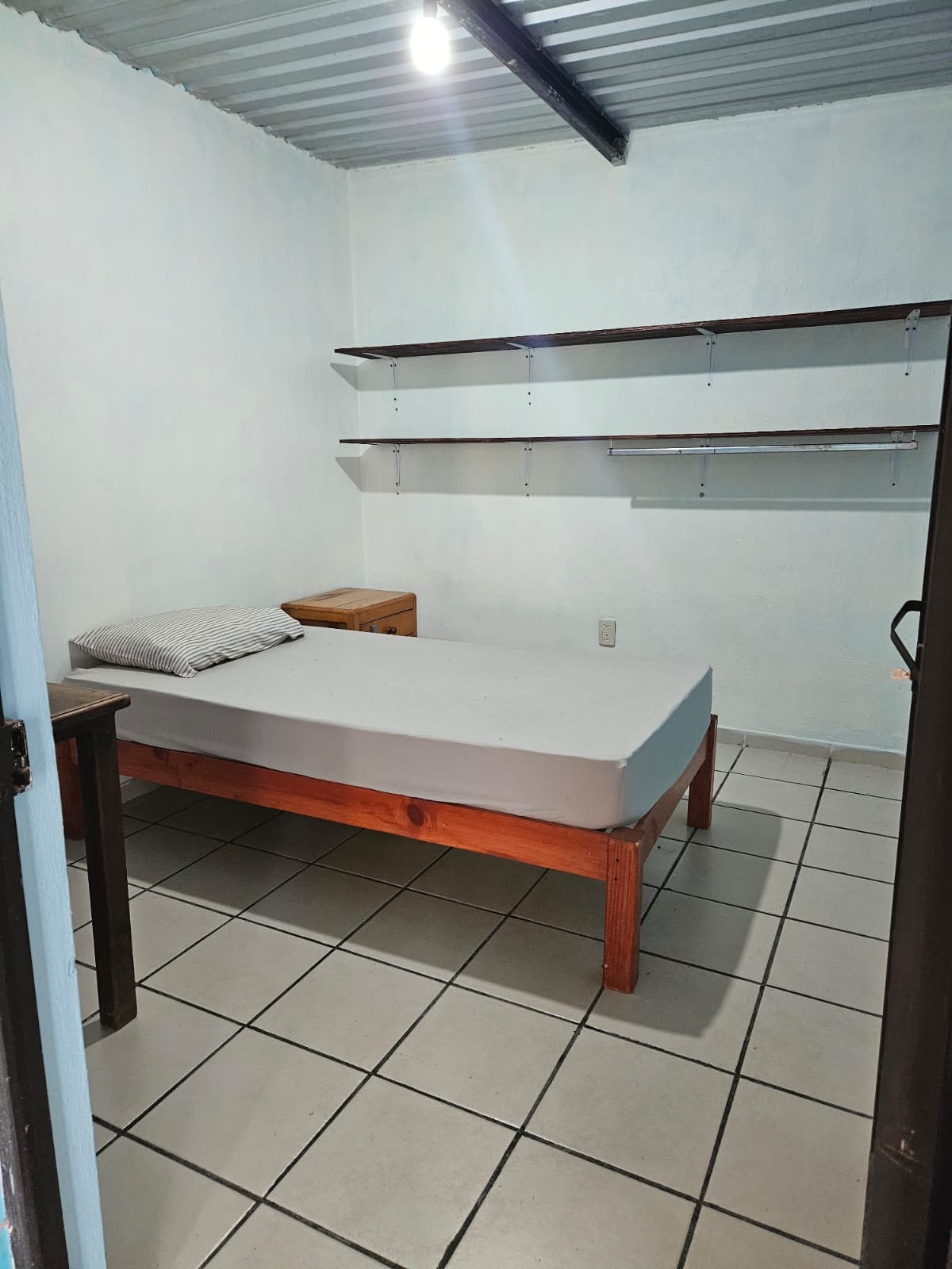 A simple sleeping area is presented with a spacious bed positioned against a light-colored wall. Minimalist wooden shelves are mounted above the bed, offering storage options, while a wooden nightstand is placed beside it. Soft lighting illuminates the room, highlighting the tiled floor.