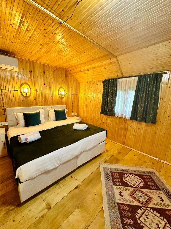 A cozy sleeping area is featured with warm wooden paneling and a sloped ceiling. A spacious bed is adorned with neutral bedding and complemented by two decorative pillows. A small rug adds texture to the wooden floor, while a window is framed by dark curtains.