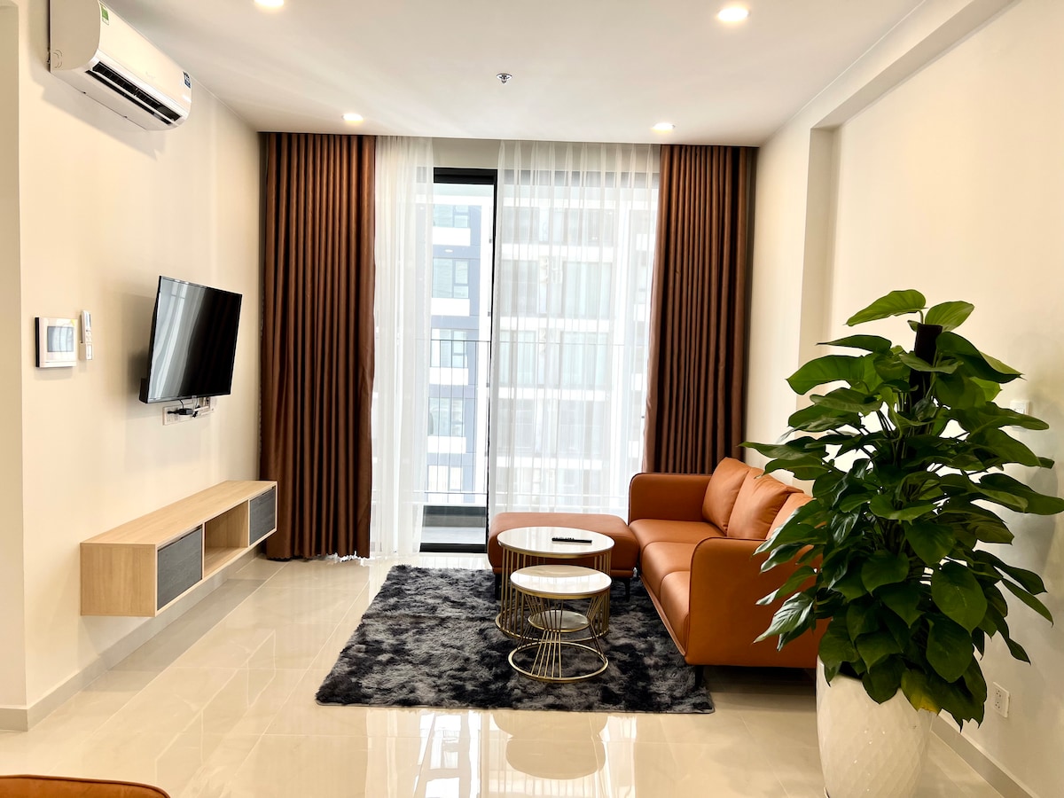 A bright living space is showcased, featuring a comfortable leather sofa paired with a circular coffee table. Natural light enters through large glass doors, which are framed by stylish curtains. A decorative plant adds a touch of greenery beside the seating area, enhancing the room's appeal.