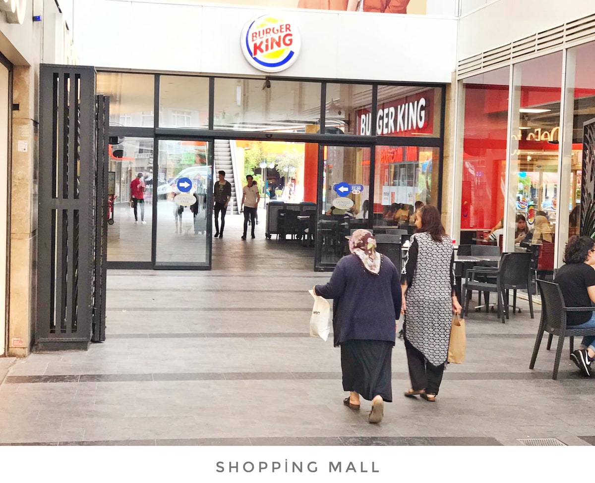 A bustling shopping mall is depicted, showcasing a wide entrance with glass doors leading to a Burger King. Two individuals are seen walking through the corridor, with various dining tables visible in the background, indicating a lively atmosphere.