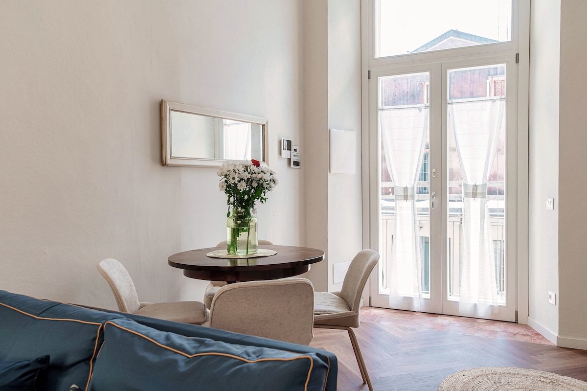 A dining area is presented with a round table surrounded by two light-colored chairs. A bouquet of flowers in a green vase adds a touch of freshness. Large windows provide ample natural light and feature sheer curtains, enhancing the bright atmosphere.