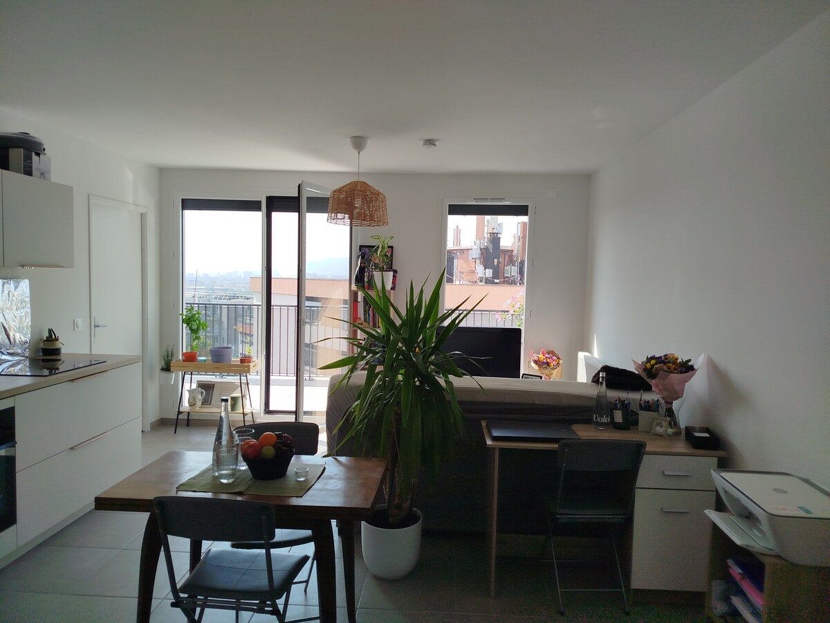 The interior space features a cozy layout with a dining table holding fresh fruit. A potted plant adds greenery. Large glass doors provide a view of the city outside. A sofa is situated near the window, creating a relaxing environment.