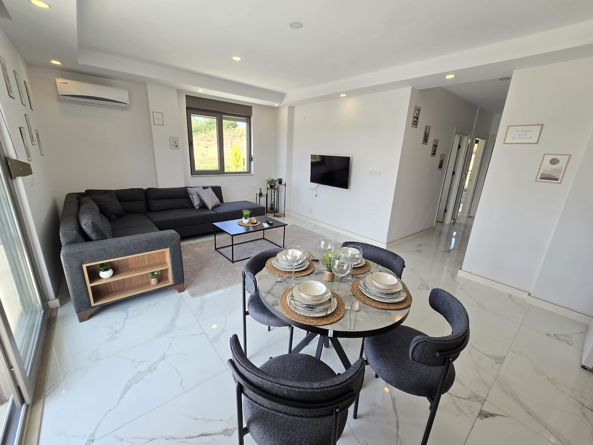 A modern living area features a sectional sofa in a neutral tone, paired with a sleek coffee table. A round dining table is set for four with simple dishware. Natural light fills the space through large windows, highlighting the polished marble floor.