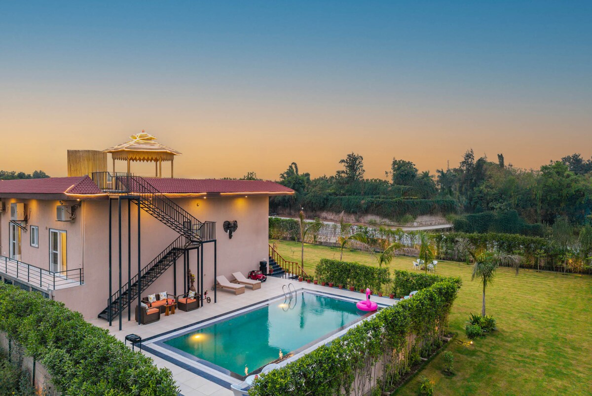 A spacious villa features a welcoming outdoor swimming pool, surrounded by manicured lawns and vibrant greenery. A wooden cabana is visible atop the villa, with sleek outdoor furniture arranged beside the pool. The scene captures a tranquil sunset, enhancing the serene atmosphere of the setting.