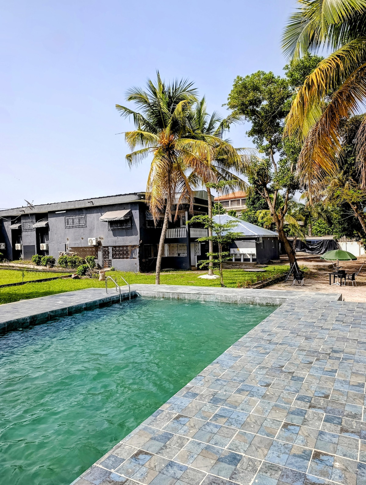 A spacious outdoor area features a clear turquoise swimming pool, surrounded by landscaped grass and palm trees. A modern two-story building with balconies is visible in the background, providing a serene setting for relaxation.