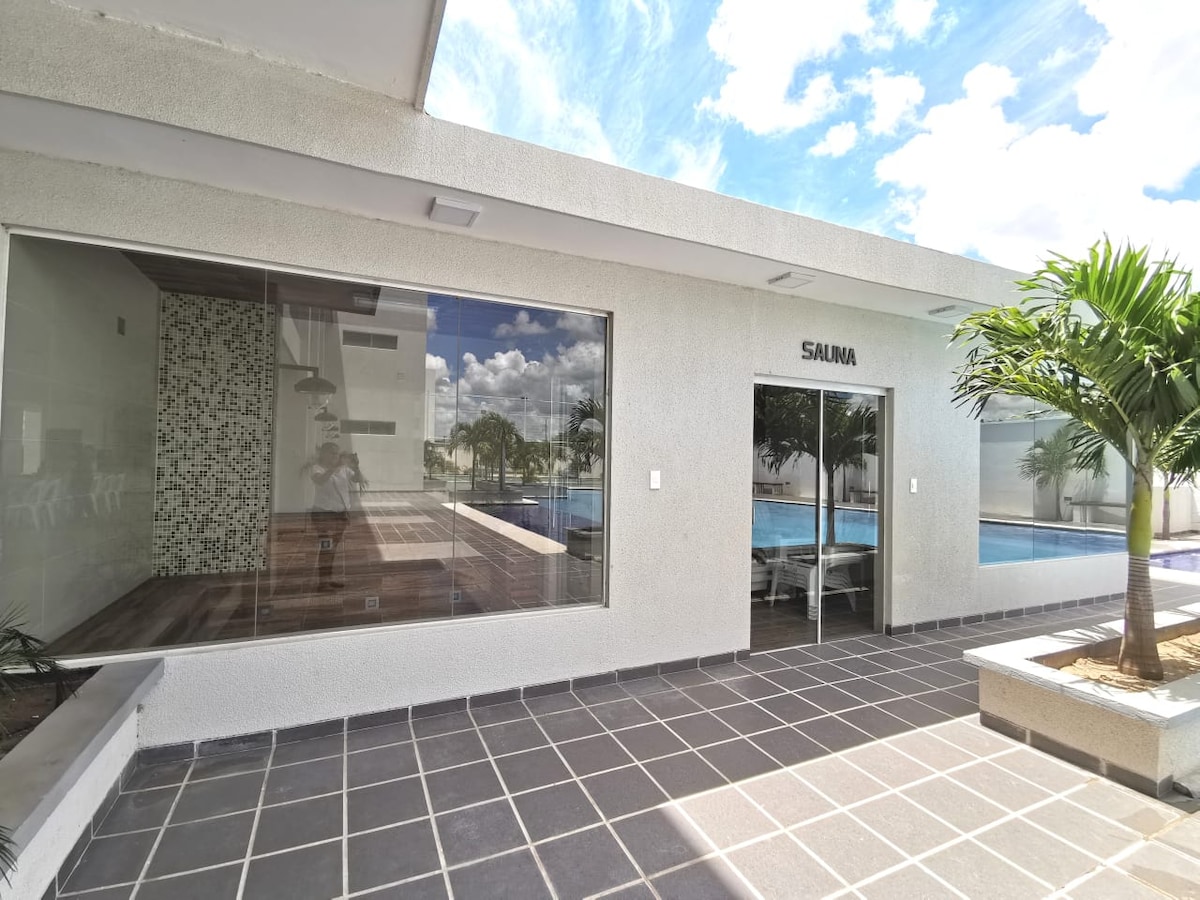 An open area is presented with large glass windows providing views of a swimming pool and outdoor space. The modern design features neutral-toned walls and tiled flooring, complemented by greenery from nearby plants, creating a bright and airy atmosphere.