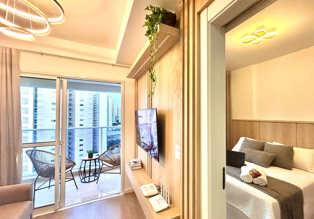 A well-lit interior showcases an integrated living area and bedroom, featuring a comfortable sofa and a Smart TV mounted on a wooden wall. An inviting balcony is visible through large glass doors, offering a view of the surrounding cityscape.