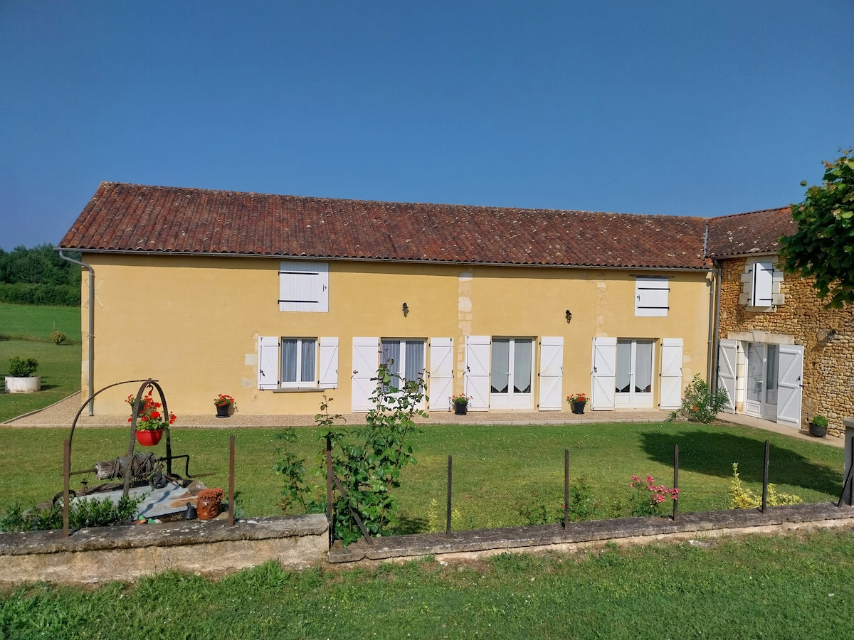 The exterior of a recently restored farmhouse is shown, featuring soft yellow walls and a sloped roof. White shutters frame several windows. A well-maintained lawn with flower pots adds a touch of greenery. A peaceful countryside landscape extends in the background.
