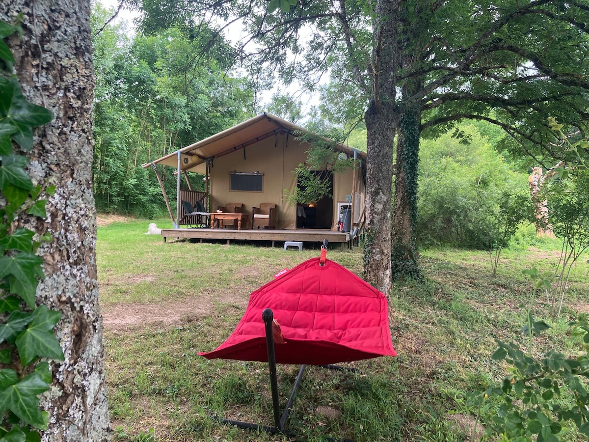 A spacious tent is positioned on a grassy area, set against a backdrop of trees. A red hammock hangs near a tree, providing a spot for relaxation. Comfortable outdoor seating is visible on the tent's deck, inviting guests to enjoy the natural surroundings.