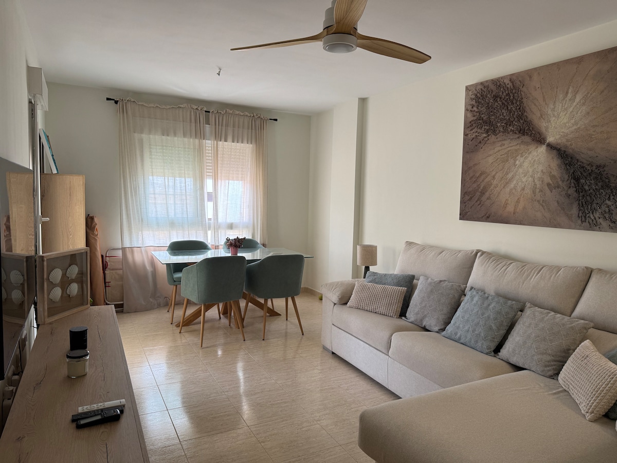 A spacious living and dining area showcases modern furnishings, including a light-colored sofa with soft cushions and a wooden dining table surrounded by teal chairs. Natural light floods in through large windows, enhancing the inviting atmosphere, complemented by a ceiling fan and minimalist wall art.