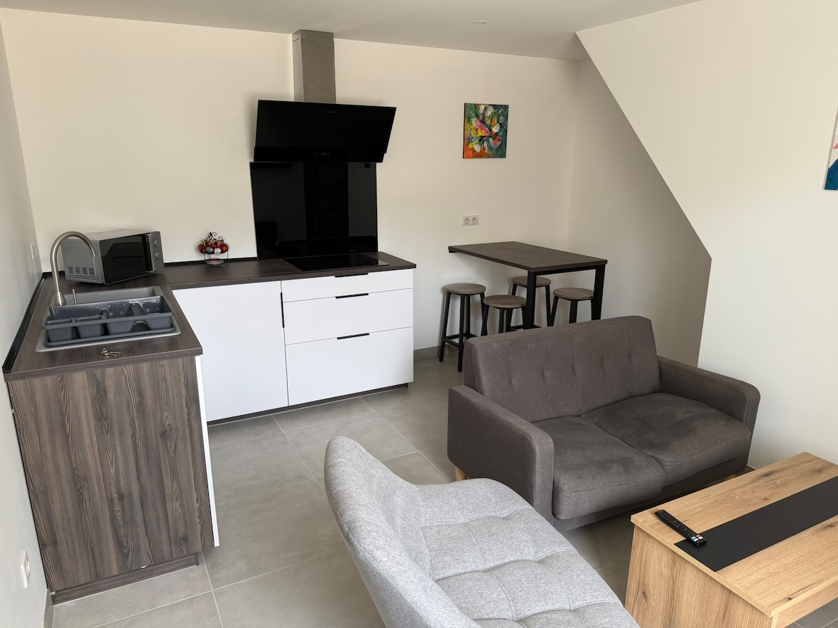 A modern open-plan living area is presented, featuring a sleek kitchen with white cabinetry and a dark countertop. A comfortable gray sofa and a light gray armchair provide seating. A small dining table with two stools is visible, alongside decorative wall art that adds a touch of color.