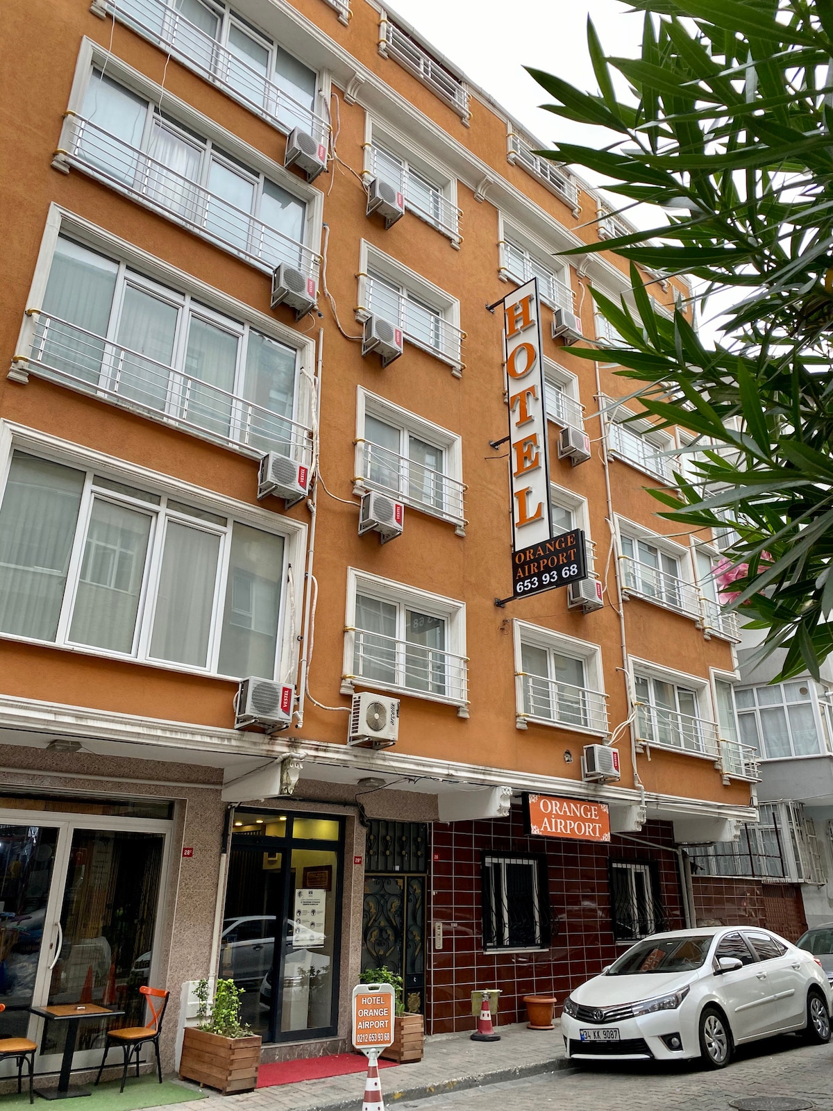 A multi-story hotel building features a vibrant orange facade with numerous air conditioning units placed along the exterior. Large windows offer ample natural light, while the entrance is marked by a sign reading 'ORANGE AIRPORT.' A parked vehicle is seen in front.