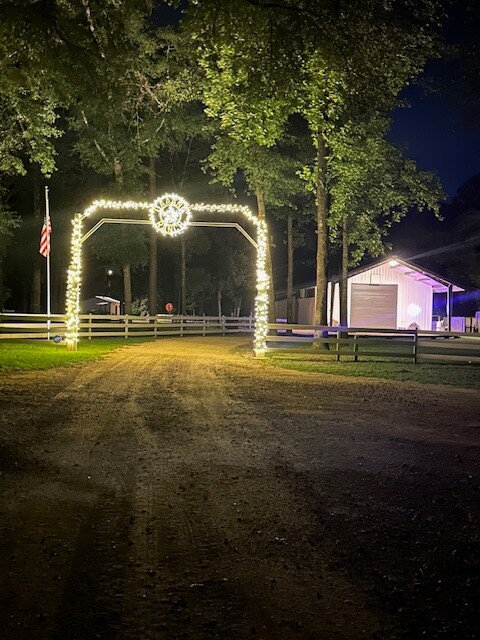 A warmly lit entrance is framed by glowing string lights, welcoming guests into a quiet forest retreat. The pathway is lined with trees, and a partially visible barn stands to the side, while an American flag gently flutters in the night breeze.