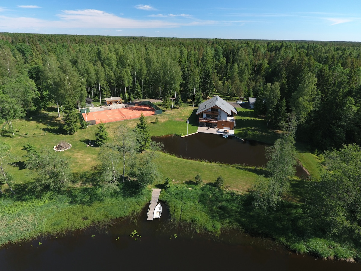 An aerial view captures a spacious property featuring a three-story house surrounded by lush greenery. A tennis court is visible, along with tranquil ponds and a small boat dock. The natural landscape suggests a serene escape into nature.