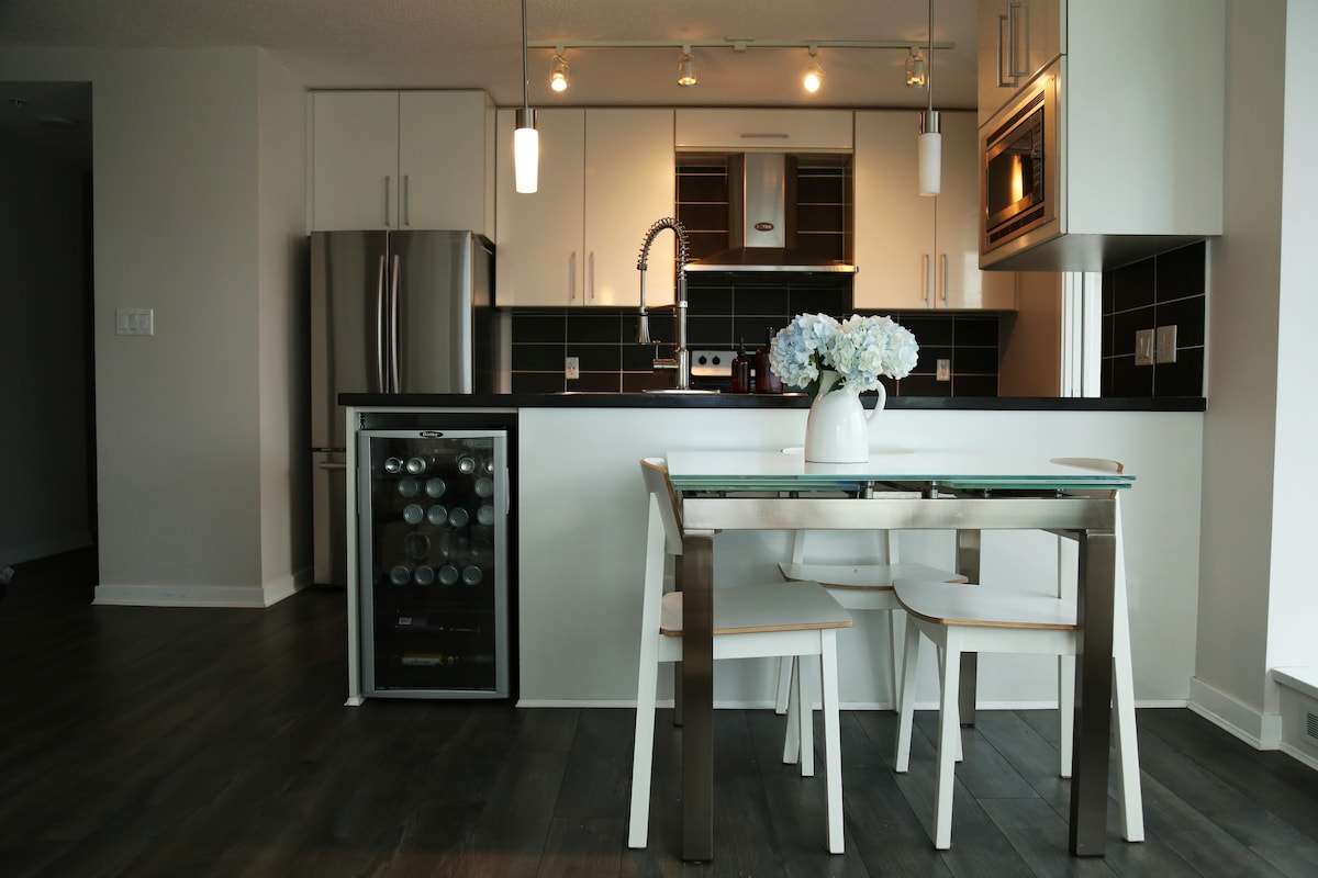 A modern kitchen area features sleek white cabinetry and stainless steel appliances. A glass dining table is accompanied by three minimalist chairs. A small wine cooler is visible, and a vase with white flowers adds a touch of elegance to the space.
