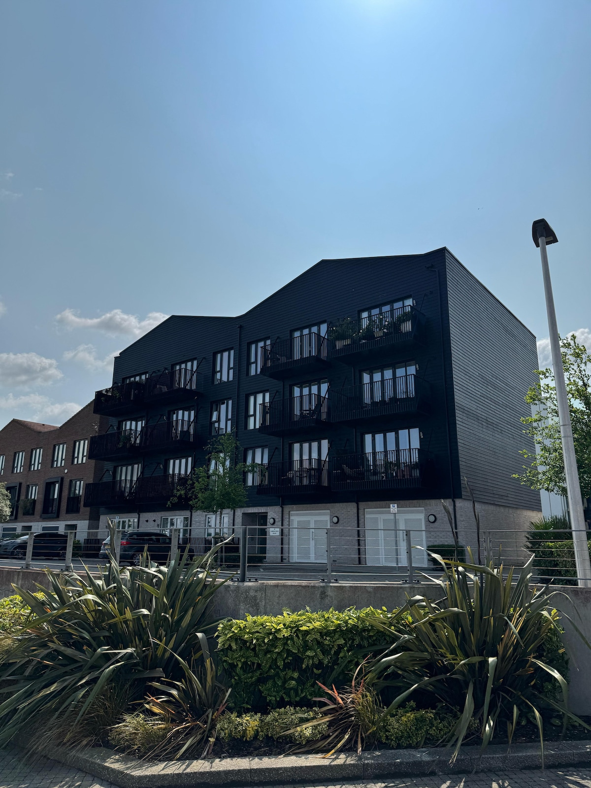 A modern three-story apartment building is depicted with a black exterior. Balconies can be seen on each level, adorned with plants. The front features a well-kept landscaped area with shrubs and palm-like plants, under a clear blue sky filled with soft clouds.