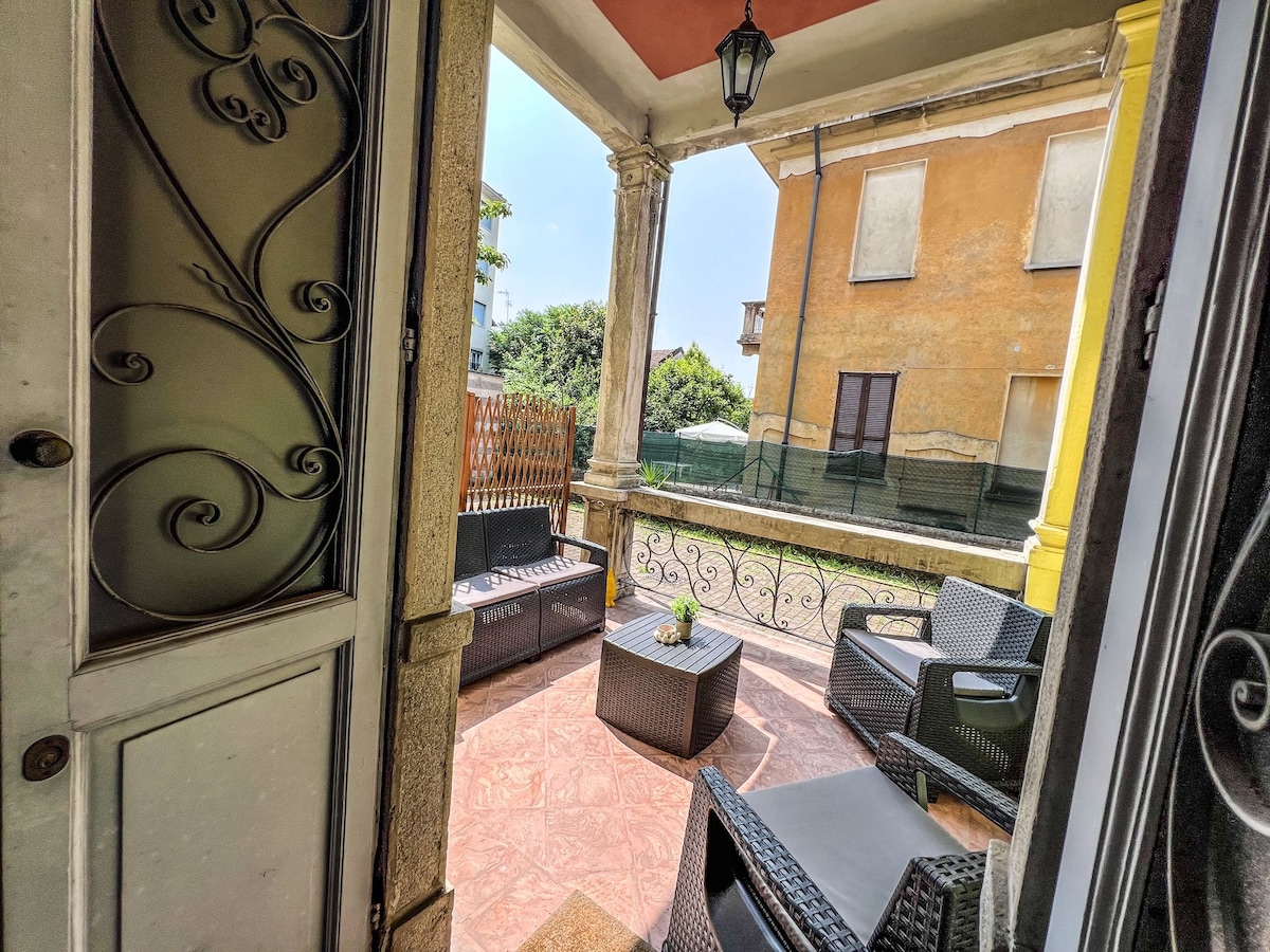 A cozy terrace area is visible through the entrance, featuring outdoor seating with dark wicker furniture and a small table. Sunlight highlights the decorative elements, creating a comfortable space for relaxation. Lush greenery is seen in the background, providing a sense of tranquility.