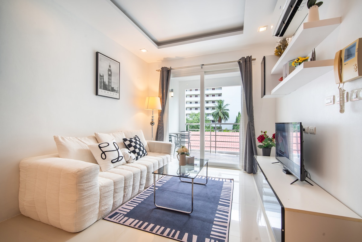 A bright living area is featured, showcasing a light-coloured sectional sofa adorned with decorative pillows. A glass coffee table sits in front of the sofa, and a television is mounted on the wall opposite. Floor-to-ceiling windows allow natural light to illuminate the space.